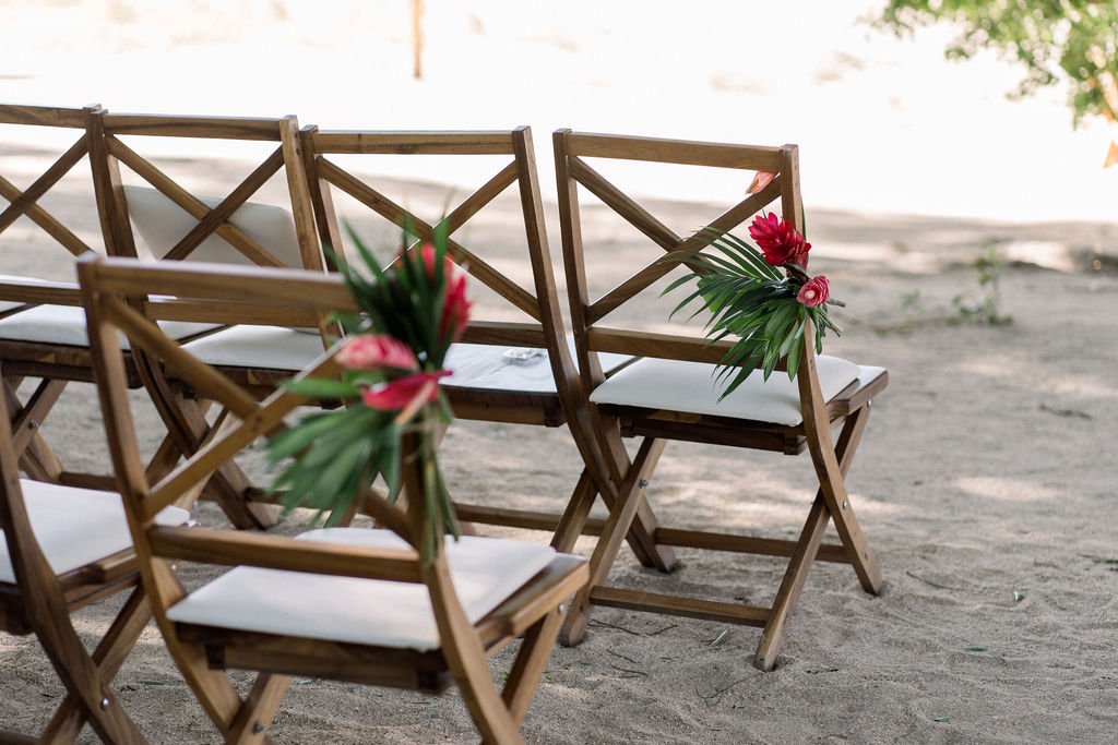 Wooden chairs with white cushions are arranged in rows on a sandy surface, each decorated with tropical flowers and green foliage.