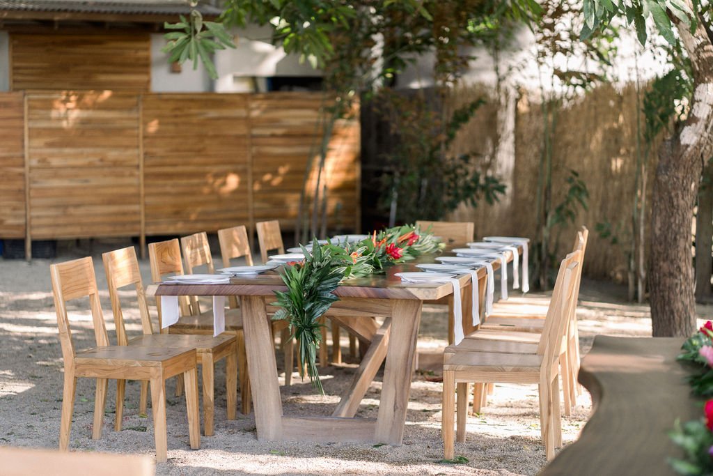 Outdoor setting with a long wooden dining table, surrounded by wooden chairs, and decorated with green and red foliage. The area is shaded by trees and has a wooden fence in the background.