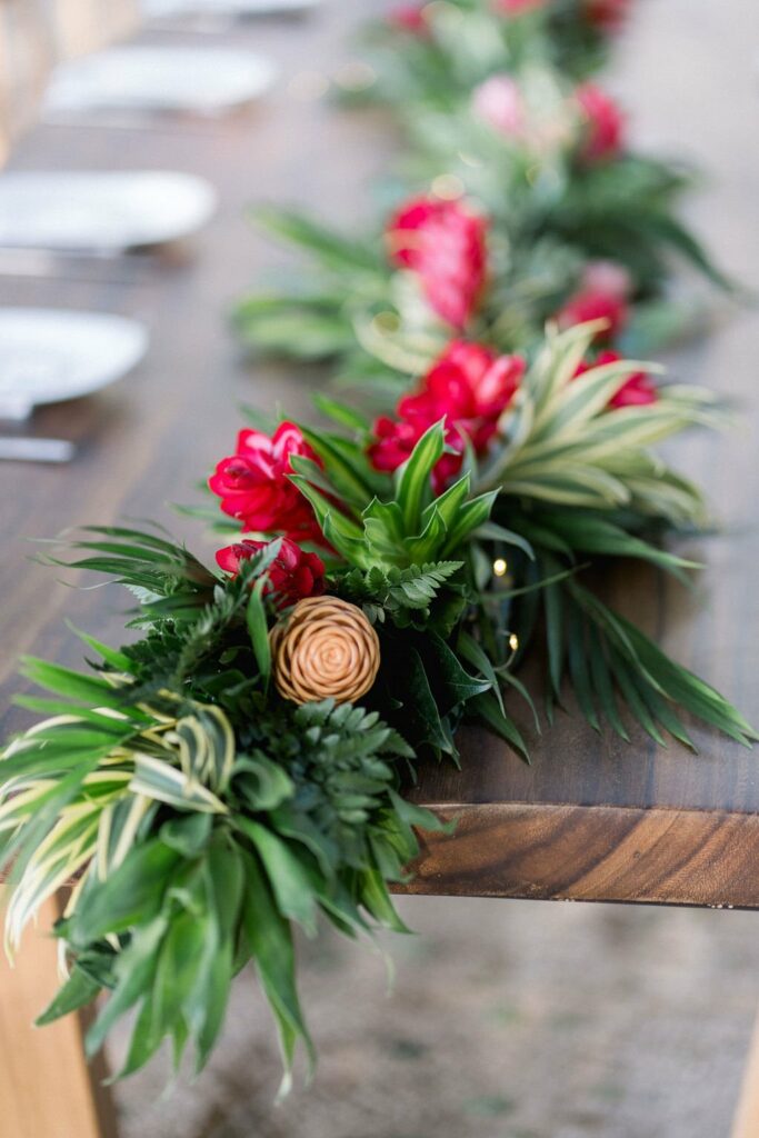 A wooden table is adorned with a lush green garland featuring red flowers and a single peach-colored rose. Plates are placed at intervals along the table.