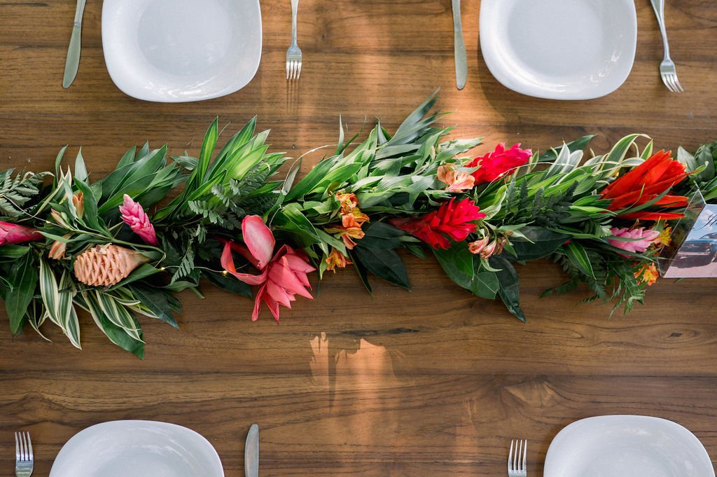 A dining table set with white plates and silverware, featuring a centerpiece of lush green foliage and vibrant red and pink flowers.