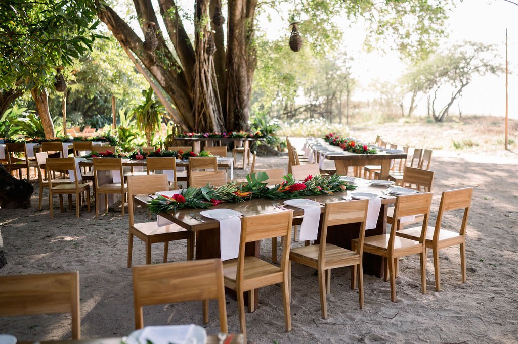 Outdoor dining area with multiple wooden tables and chairs set up under large trees. Each table is decorated with a white table runner and greenery arrangements.