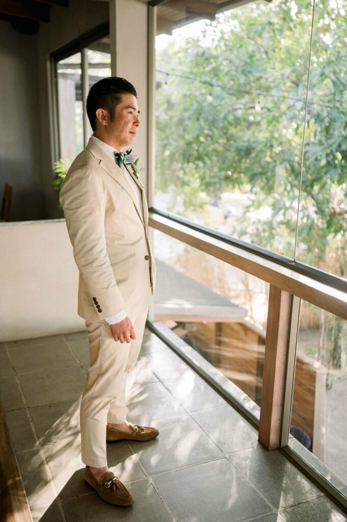 A man in a beige suit stands indoors by a large window, looking outside. Natural light illuminates the space.