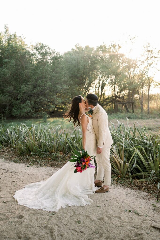 A bride and groom share a kiss outdoors with greenery and sunlight in the background. The bride holds a colorful bouquet and wears a white dress, while the groom is in a light-colored suit.