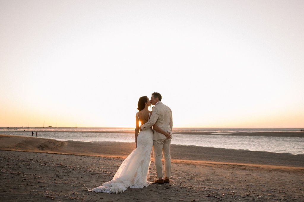 A couple in wedding attire stands embraced on a beach at sunset, facing the ocean. The bride wears a white gown, and the groom is in a light-colored suit. The sky is clear with the sun near the horizon.