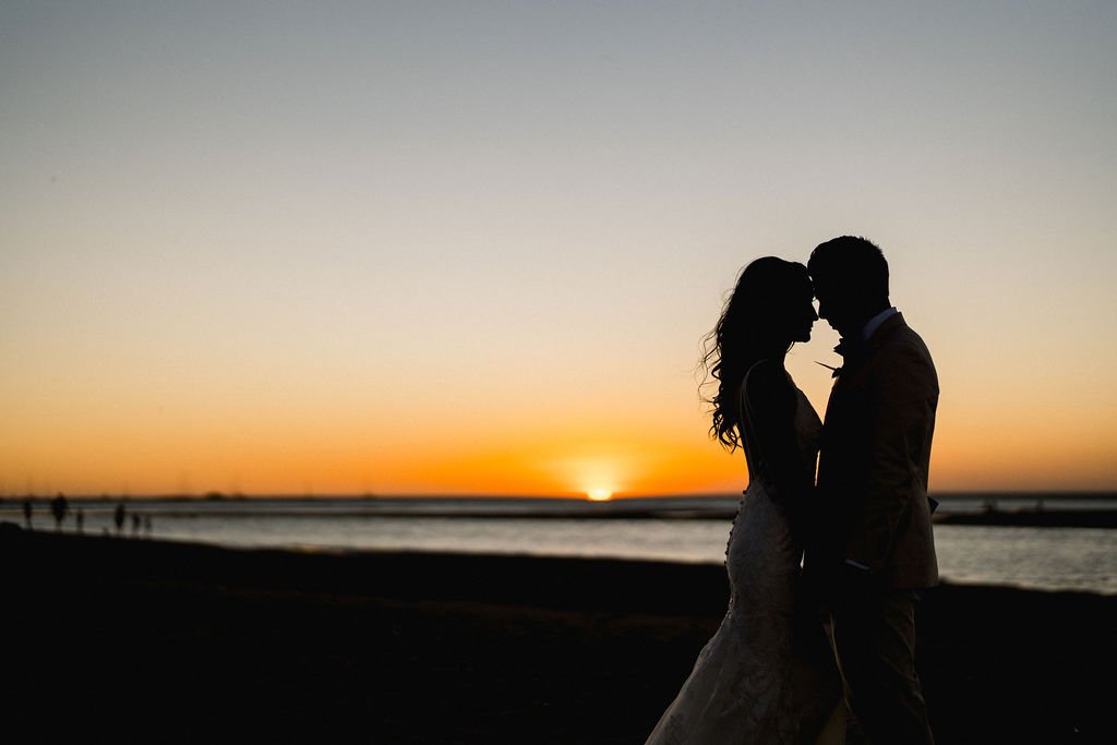 A silhouetted couple stands close together on a beach at sunset, with the sun low on the horizon over calm waters.