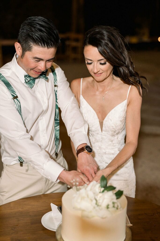 A bride and groom cut a white frosted wedding cake together. The groom wears a white shirt with green suspenders and a bow tie, while the bride wears a white lace dress.