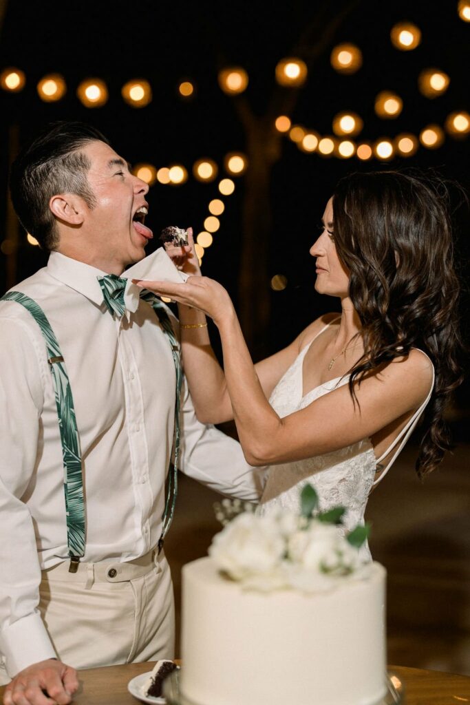 A woman in a white dress feeds cake to a man in a white shirt and suspenders, with string lights in the background at a nighttime event.