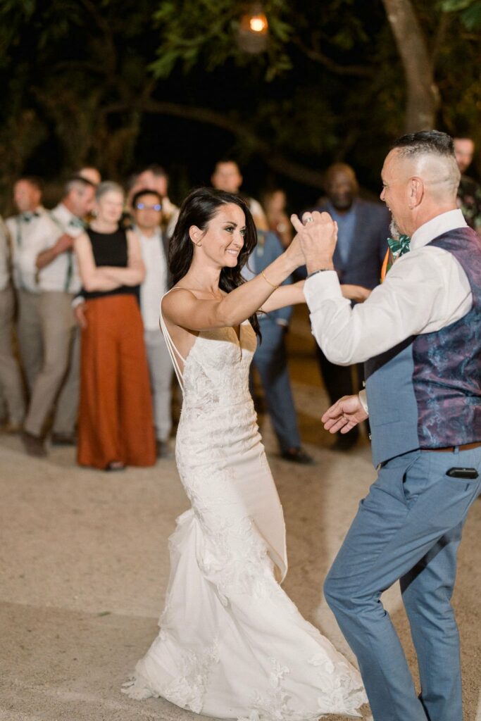 A bride in a white dress and a man in a blue suit and vest are dancing outdoors at a wedding reception, with a group of people watching and smiling in the background.
