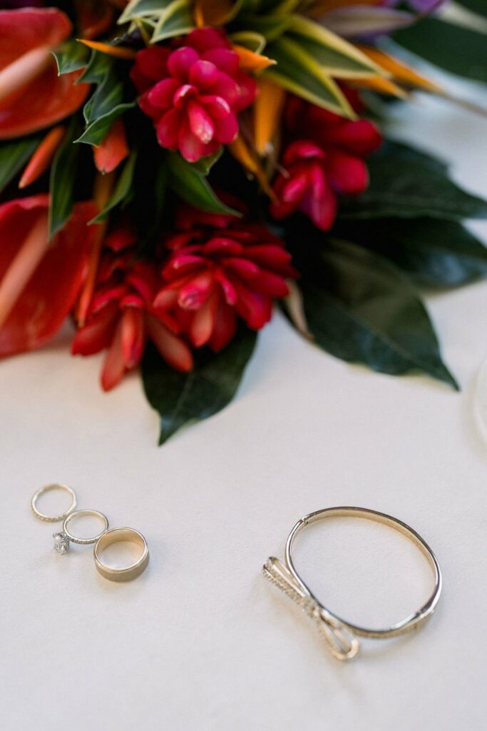 A close-up of two gold rings, three silver rings, and a bracelet on a white surface next to a floral arrangement with red and pink flowers.