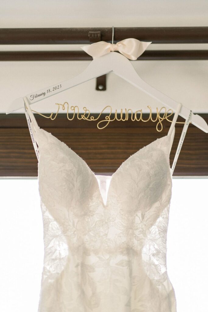 A white lace wedding dress on a custom hanger that reads "Mrs. Juneau," dated February 18, 2023.