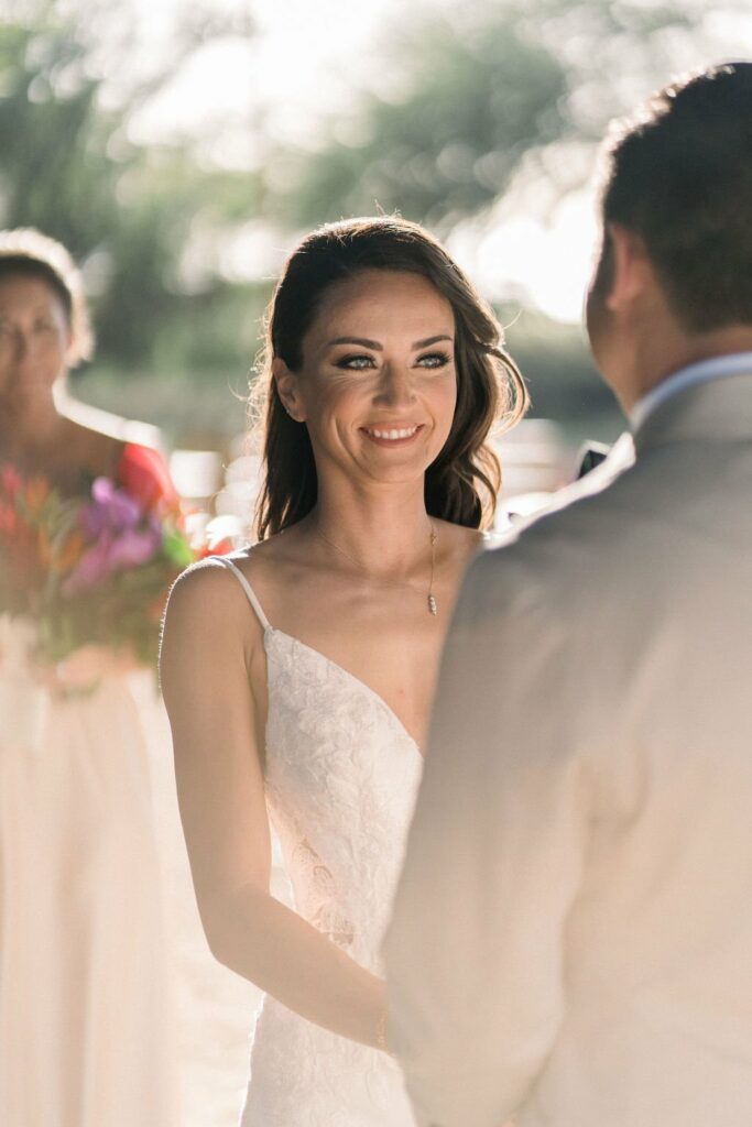 A bride in a white dress smiles as she stands next to a man in a suit during a wedding ceremony. Another person in the background holds colorful flowers.