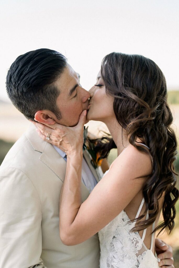 A bride and groom share a kiss on their wedding day, with the bride holding the groom's face and their eyes closed. The groom is wearing a light-colored suit, while the bride is in a white dress.