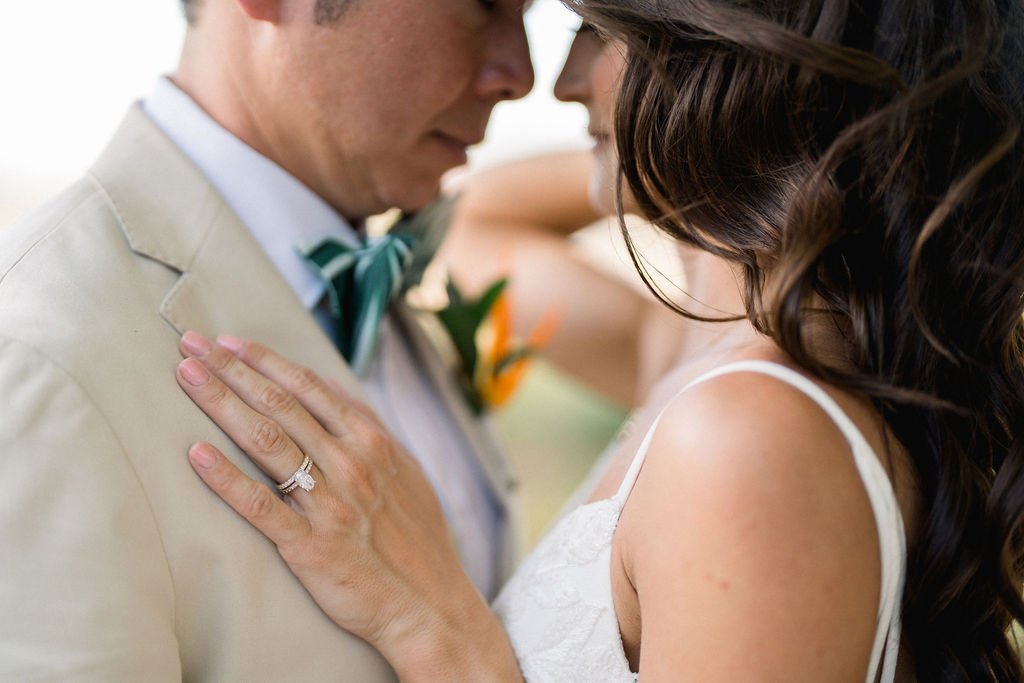 A close-up of a couple embracing, with the woman's hand showing a wedding ring placed on the man's chest, dressed in light-colored formal attire with a teal bow tie.