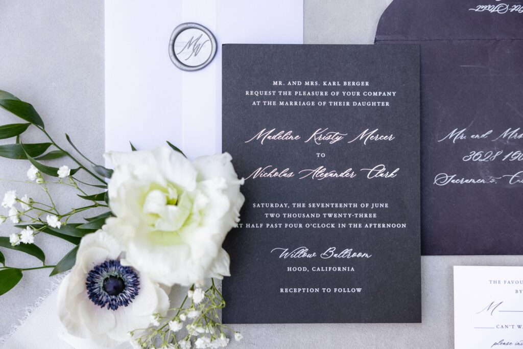 A wedding invitation with white and gold text on a black card, surrounded by white flowers and an envelope, announces the marriage of Madeline Kristy Mercer and Nicholas Alexander Teir.