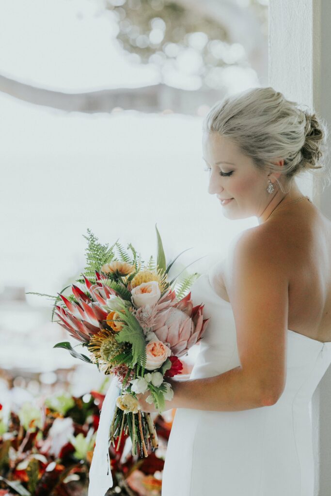 A bride in a white dress stands holding a colorful bouquet of flowers, looking down with a smile.