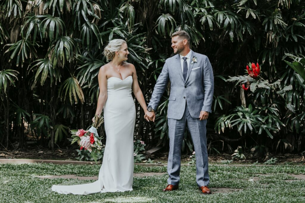 A bride in a white strapless gown and a groom in a light gray suit hold hands while standing outdoors in front of lush greenery.