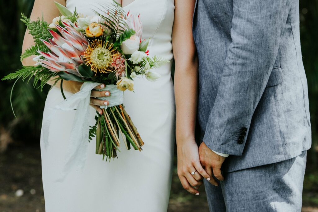 A bride in a white dress and groom in a gray suit stand side by side, holding hands. The bride holds a bouquet of flowers.