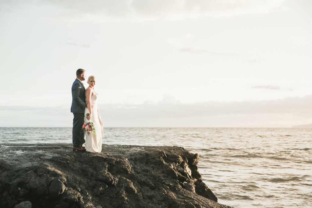 A couple in wedding attire stands on a rocky shore overlooking the ocean at sunset. The bride holds a bouquet of flowers.