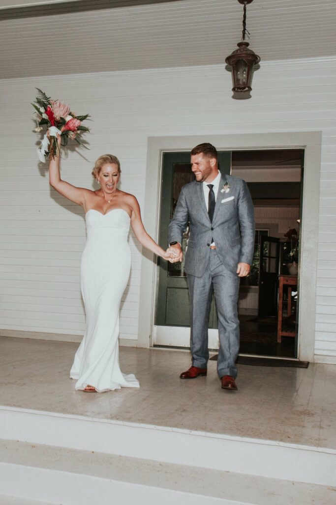 A bride in a white strapless gown holding a bouquet raises her arm, standing beside the groom in a gray suit holding her hand, both smiling and stepping down from a porch.