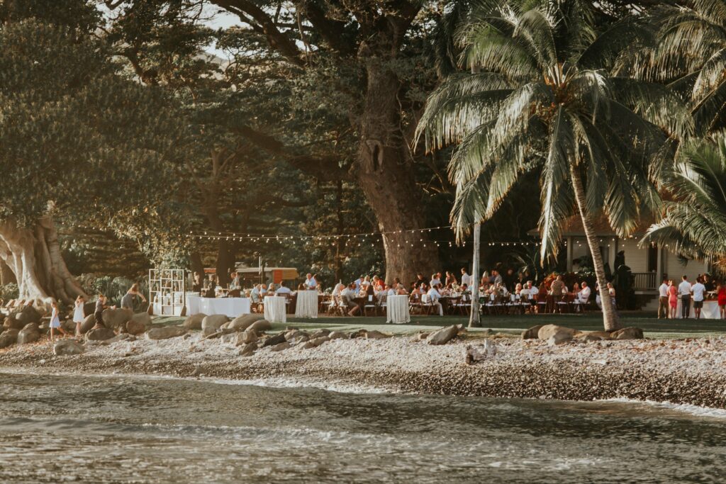 A waterfront outdoor event with numerous guests seated at tables under string lights, surrounded by lush tropical trees and palm trees, beside a pebbled shoreline.