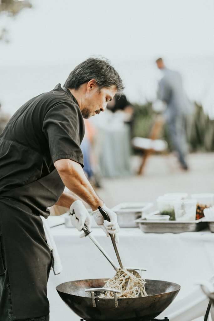 A chef wearing a black apron and gloves cooks noodles in a large wok at an outdoor event. In the background, tables and other attendees are visible.