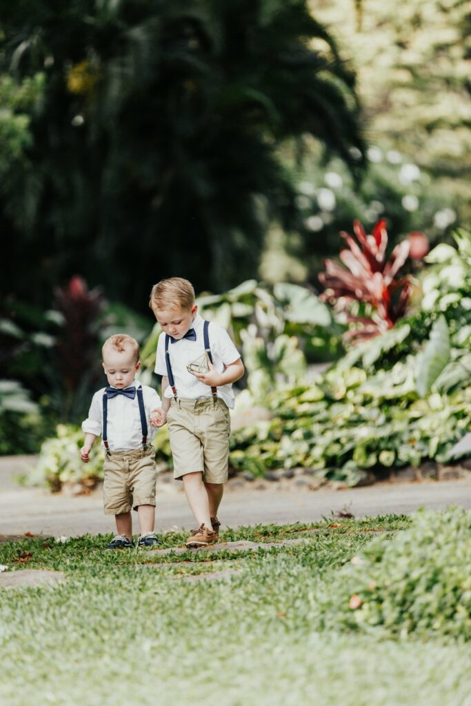Two young boys, dressed in matching beige shorts and white shirts with suspenders and bow ties, walk hand-in-hand on a garden path surrounded by lush greenery.