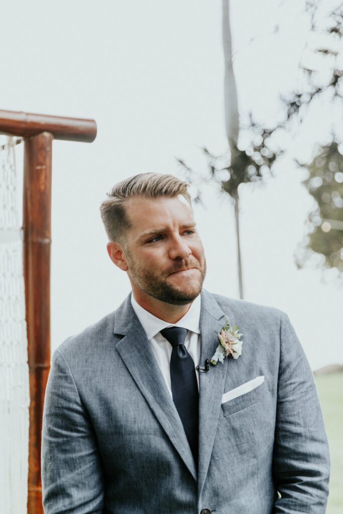 A man in a grey suit with a white shirt and black tie stands outdoors, wearing a small boutonniere on his left lapel.