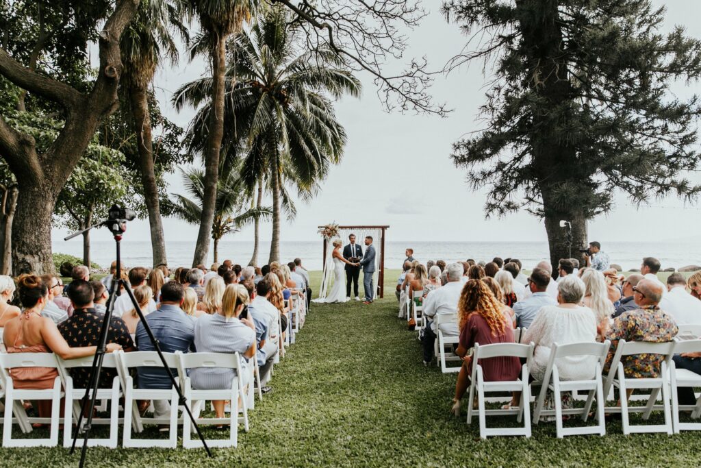 A wedding ceremony is taking place outdoors. Guests are seated on either side of an aisle lined with palm trees, leading to a couple standing under a wooden altar by the ocean.