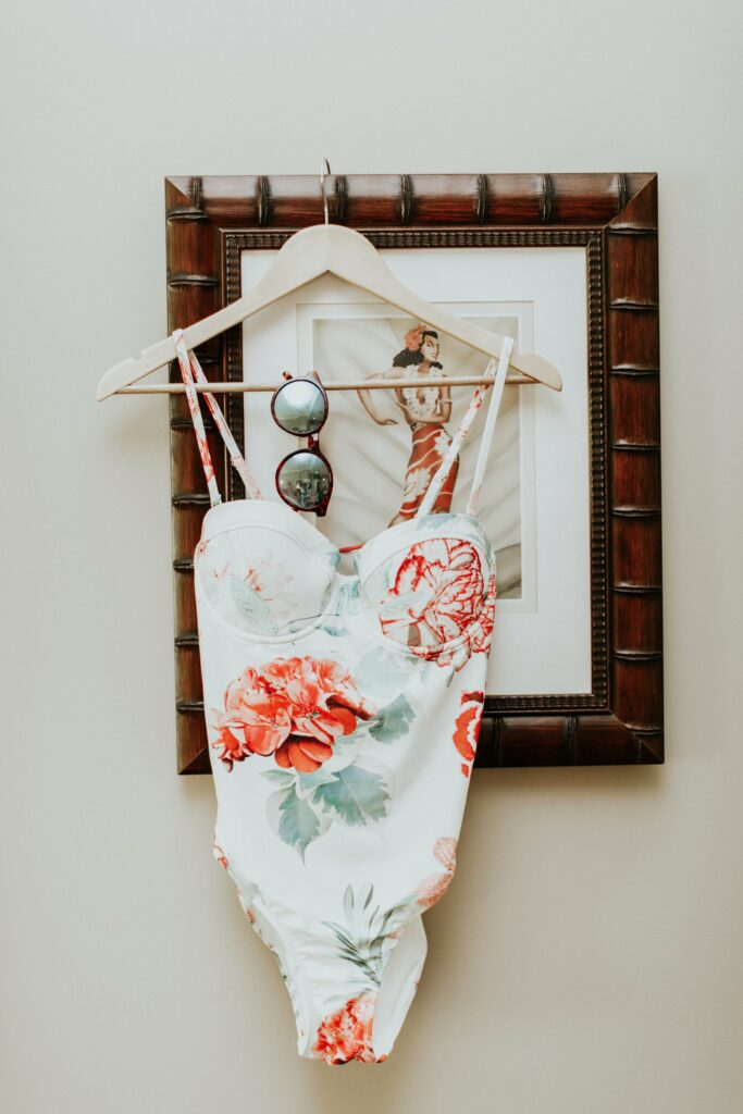 A floral-patterned swimsuit and a pair of sunglasses hang on a wooden hanger, which is placed over a framed picture of a woman in a dress.