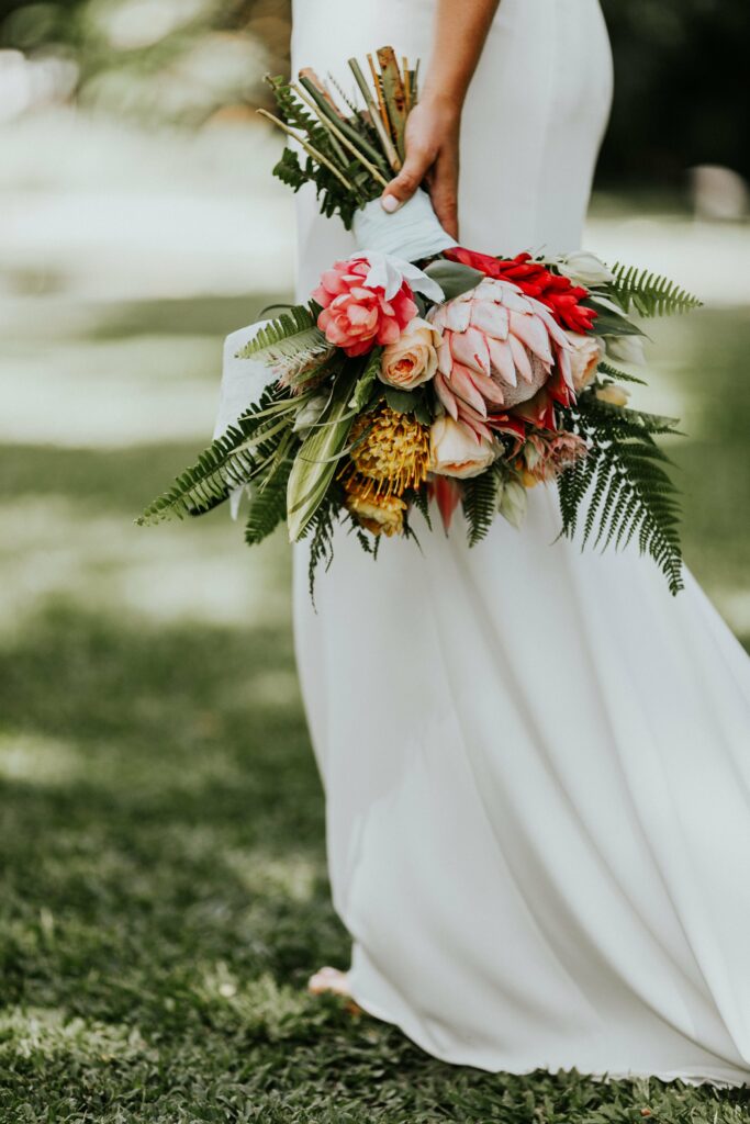 A person in a white dress holds a bouquet of brightly colored flowers and greenery while standing on grass.