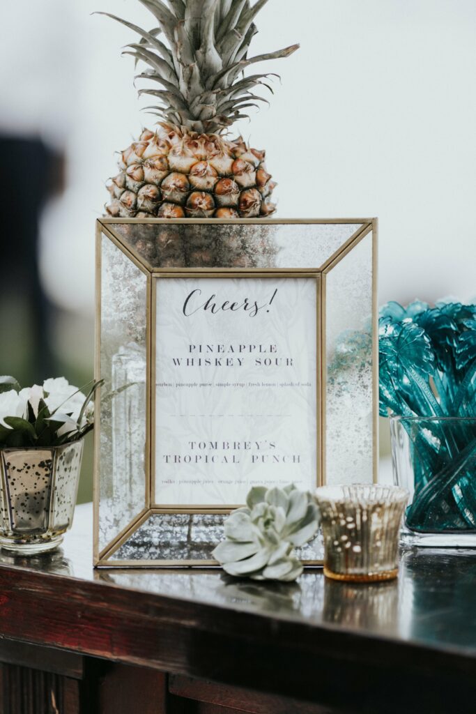 A framed menu on a table lists drink options, topped with a pineapple. The table holds various small plants and glass candle holders, with green stirrers to the right.
