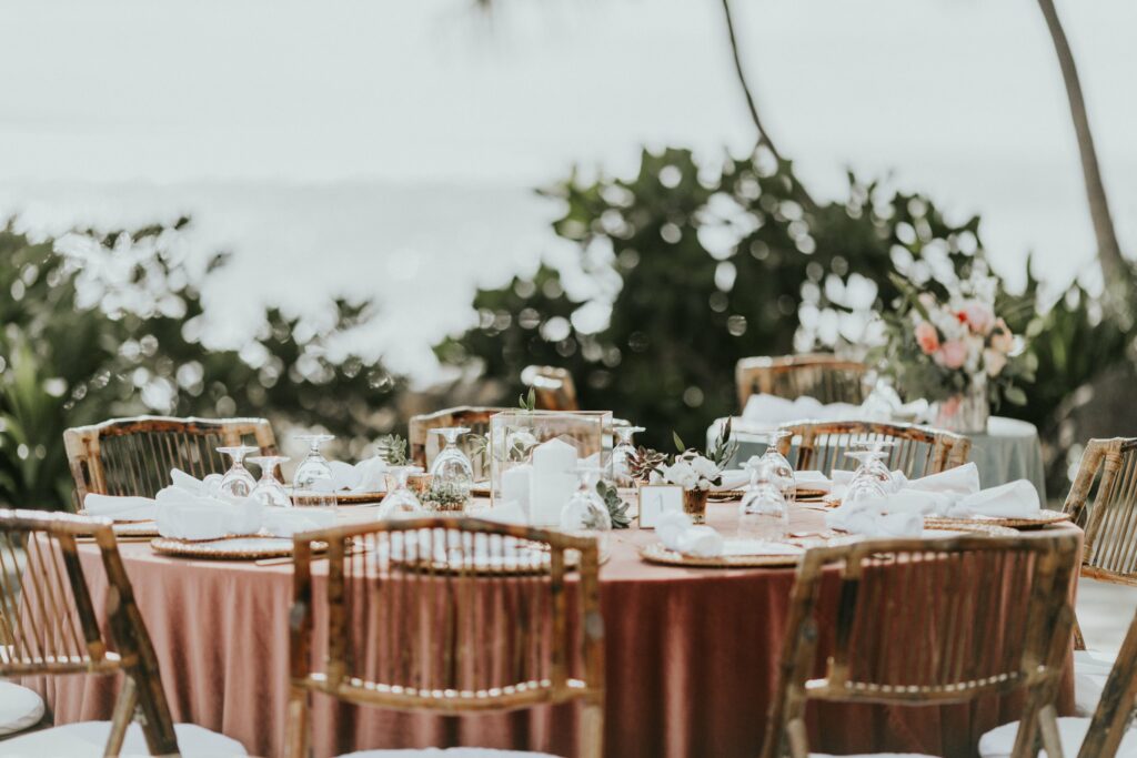 An outdoor dining setup with round tables draped in red tablecloths, wooden chairs, neatly folded napkins, glassware, and floral centerpieces amidst a natural, lush background.