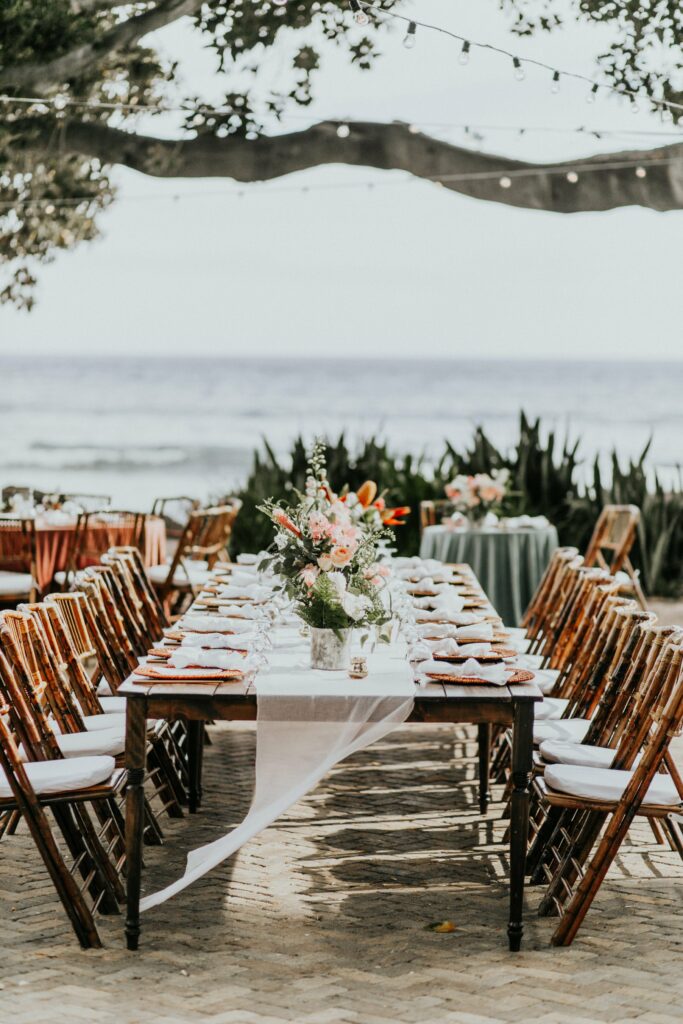 An elegantly set outdoor dining table on a beach, adorned with floral centerpieces and surrounded by wooden chairs. String lights hang above, and the ocean is visible in the background.