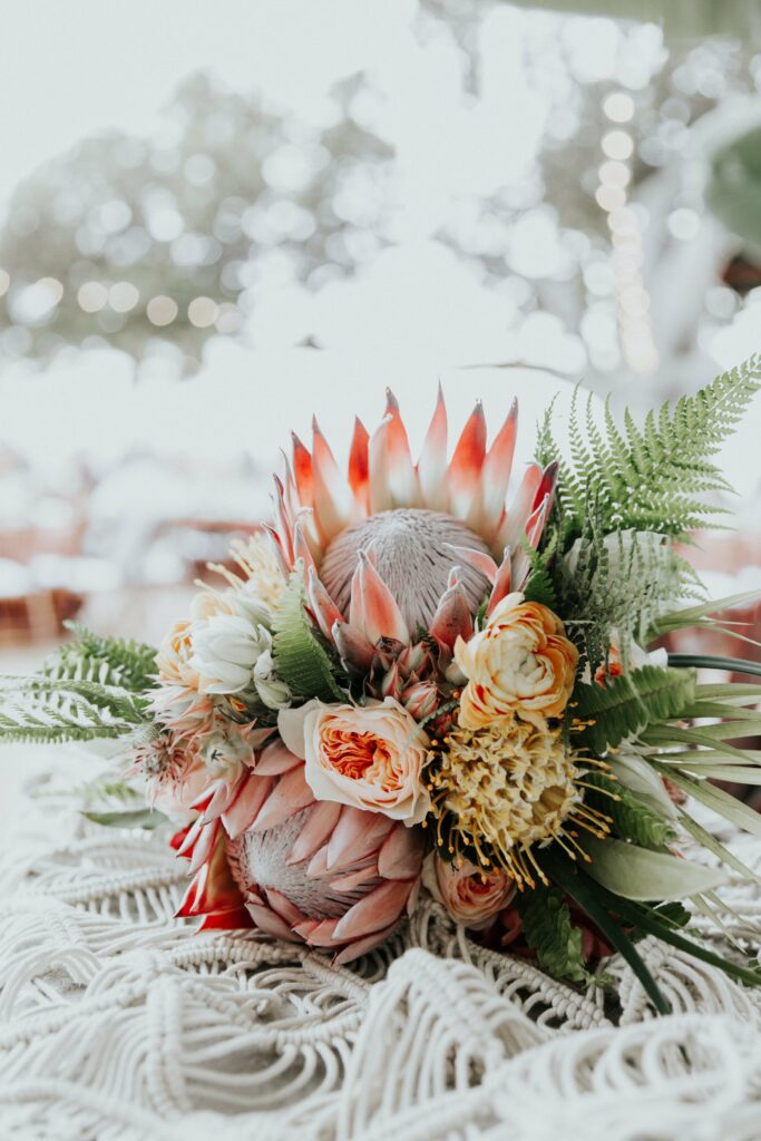 A bouquet featuring a large protea flower, surrounded by various flowers and greenery, is placed on a white lace surface.