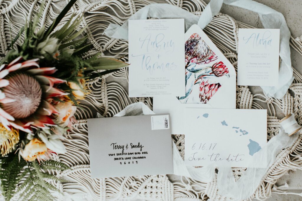 A collection of wedding stationery including invitations, envelopes, and save-the-date cards are arranged on a macrame surface, alongside a floral bouquet and ribbon.