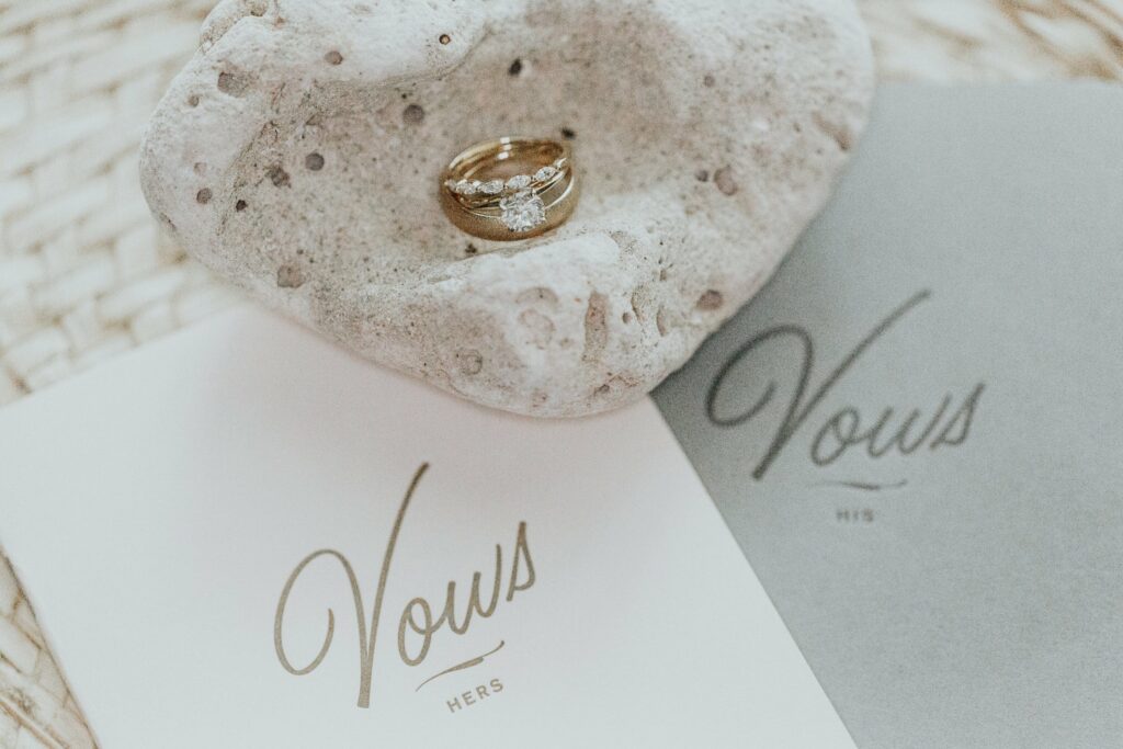 A gold ring with a diamond is placed on a stone, surrounded by two vow booklets labeled "Hers" and "His".