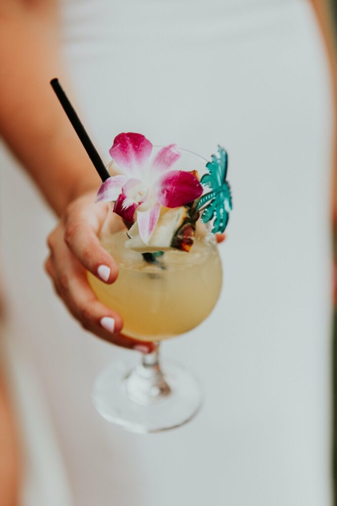 A person holding a tropical cocktail garnished with a flower, pineapple slice, and a decorative green element.
