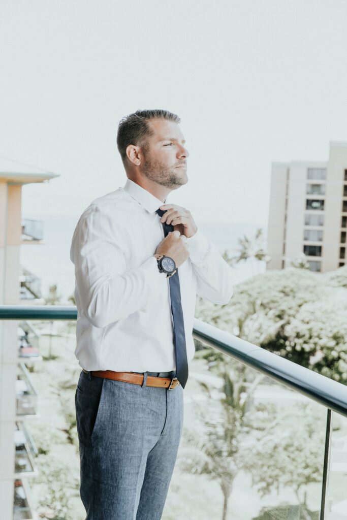 A man in a white shirt and blue tie adjusts his necktie while standing on a balcony overlooking a cityscape with buildings and greenery in the background.