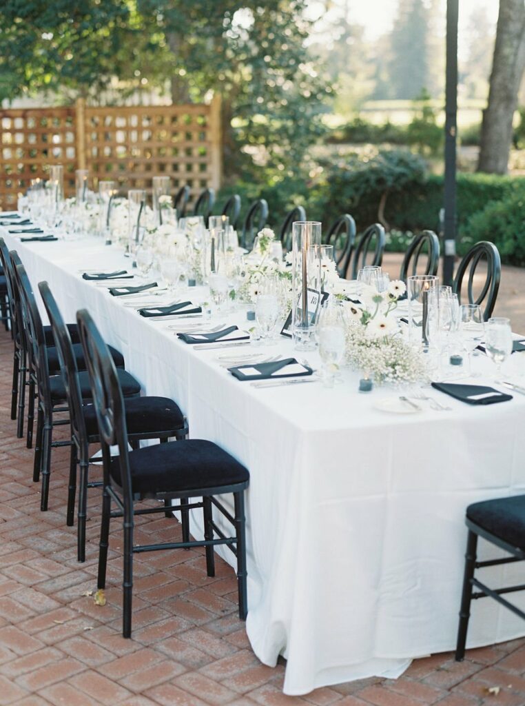 A long outdoor dining table is set with white tablecloth, black chairs, glassware, candles, and white floral centerpieces. The table is situated on a brick patio with a wooden lattice in the background.