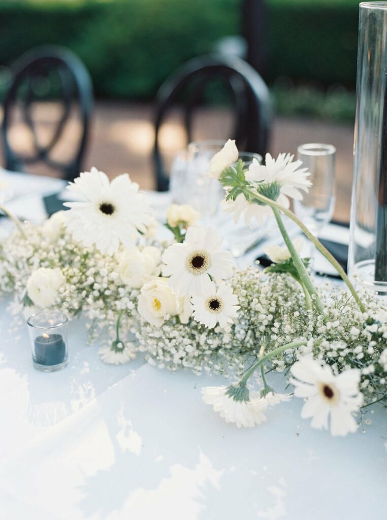 A table is decorated with a white floral arrangement, including daisies and baby’s breath. There are candles and glassware set alongside the arrangement.