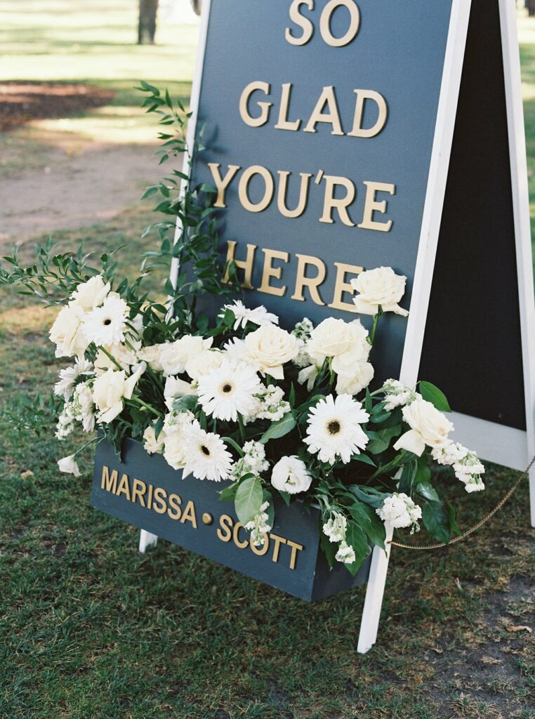 A signboard with gold text reads "SO GLAD YOU'RE HERE," with white flowers at the bottom and the names "MARISSA • SCOTT" displayed below the flowers. It is set outdoors.