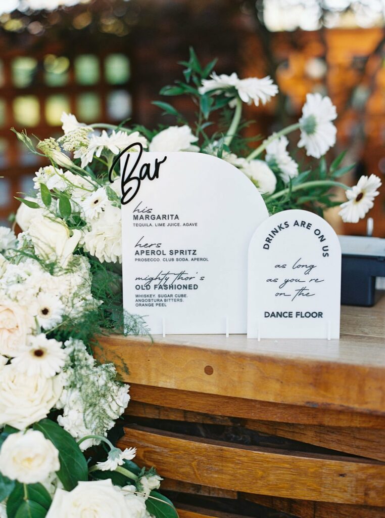 A bar menu with drink options stands amidst white flowers. It lists Margarita, Aperol Spritz, and Old Fashioned as available cocktails. A sign next to it reads, "DRINKS ARE ON US as long as you are on the DANCE FLOOR.