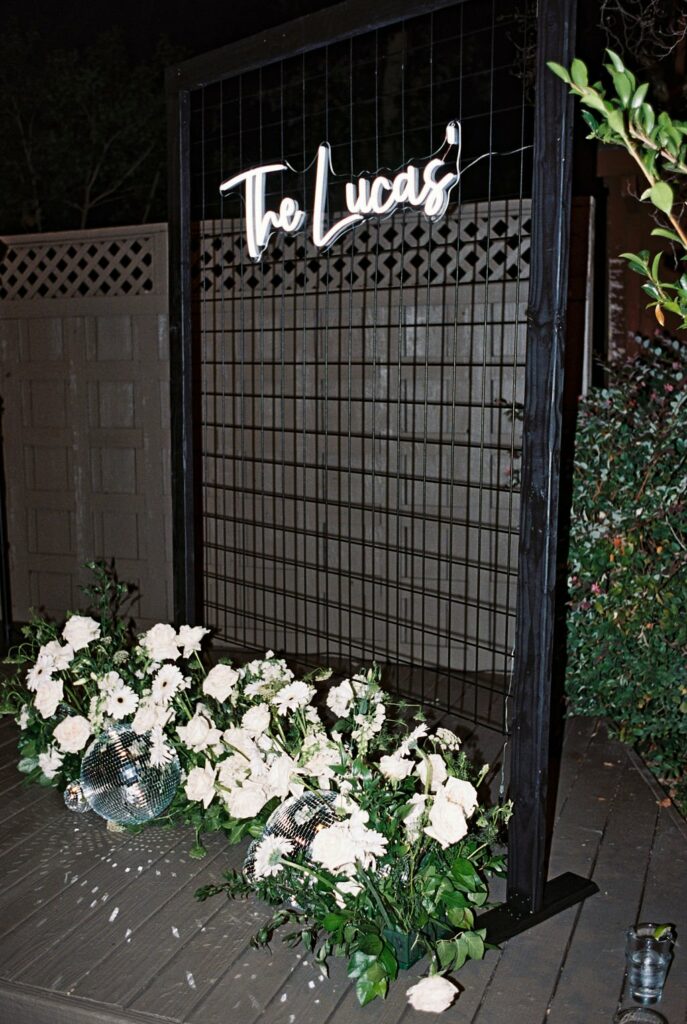 A wire frame display with a sign reading "The Lucas'" is adorned with white roses and greenery at its base. Two disco balls are also among the flowers. A wooden lattice fence is in the background.