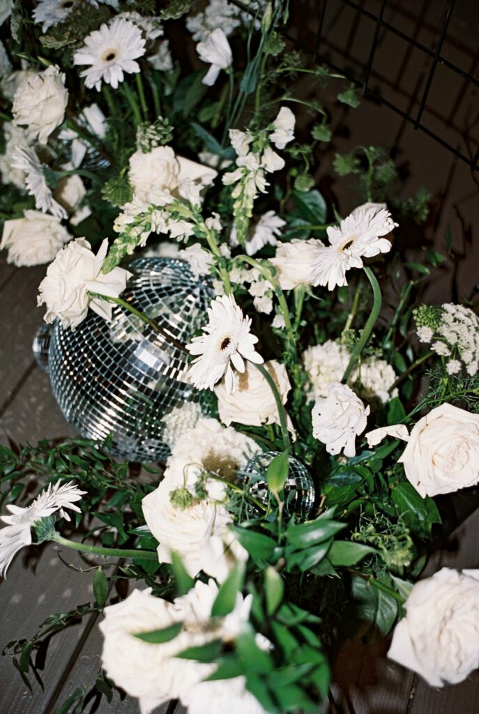 A display of white flowers and green foliage surrounds a mirrored disco ball on a wooden surface.