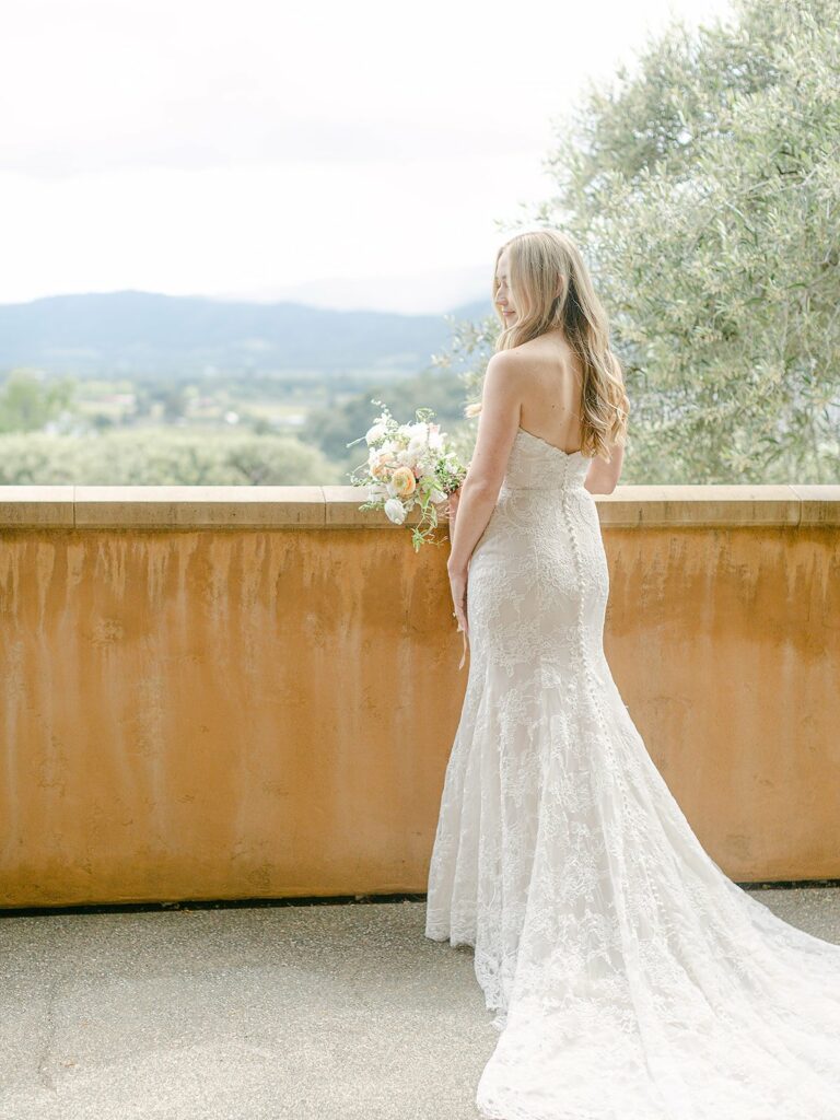 A bride in a white lace wedding dress stands with her back to the camera, holding a bouquet and looking over a stone railing with a scenic landscape in the background.