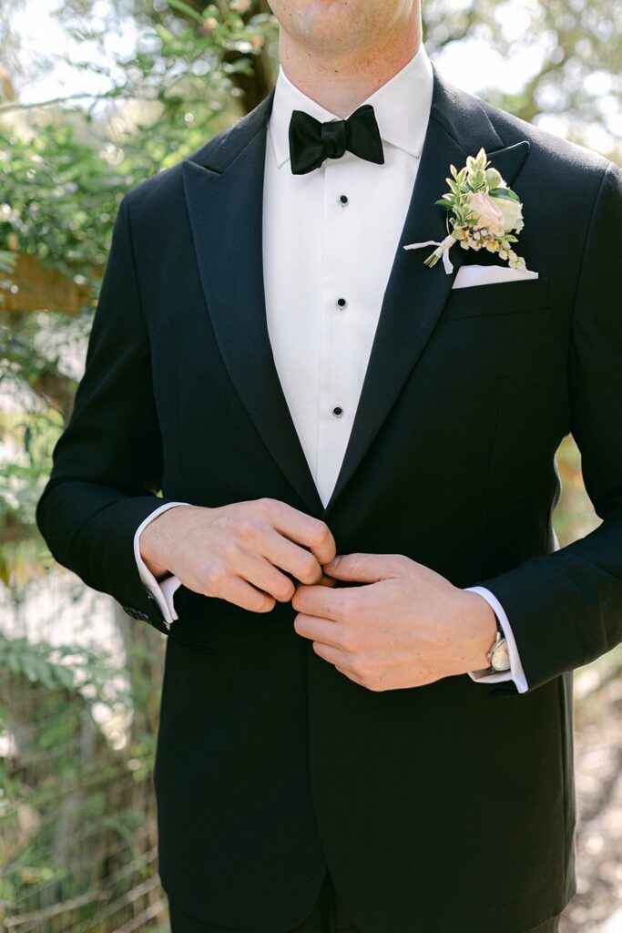 A person in a black tuxedo with a bow tie and boutonniere adjusts their jacket outdoors.