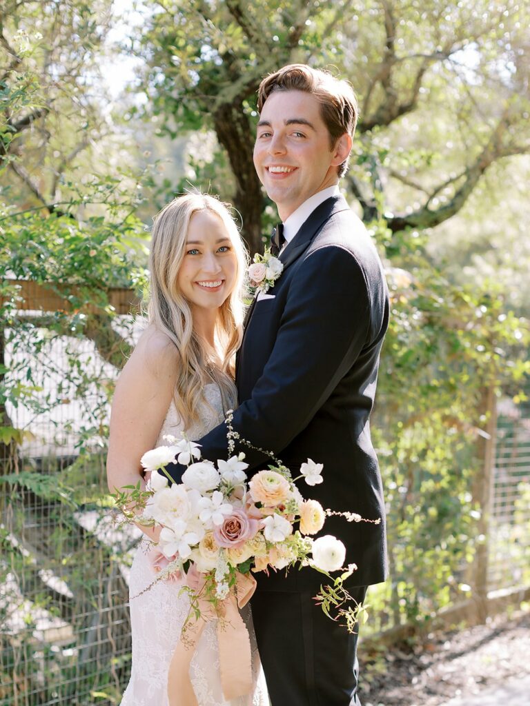 A couple dressed in wedding attire stands outdoors, smiling. The woman holds a bouquet of white and pink flowers while the man has his arm around her. Greenery is visible in the background.
