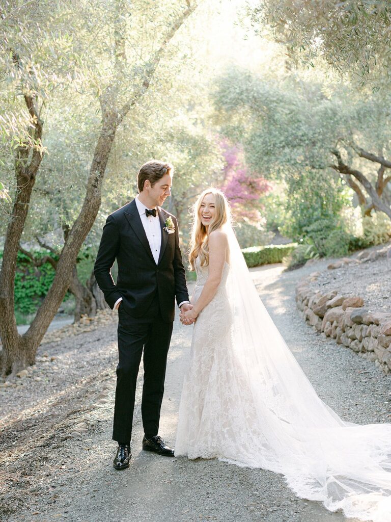 A couple in wedding attire stand hand-in-hand on a tree-lined path. The bride wears a lace gown with a long veil, and the groom is in a black tuxedo. They are outdoors with sunlight filtering through the trees.