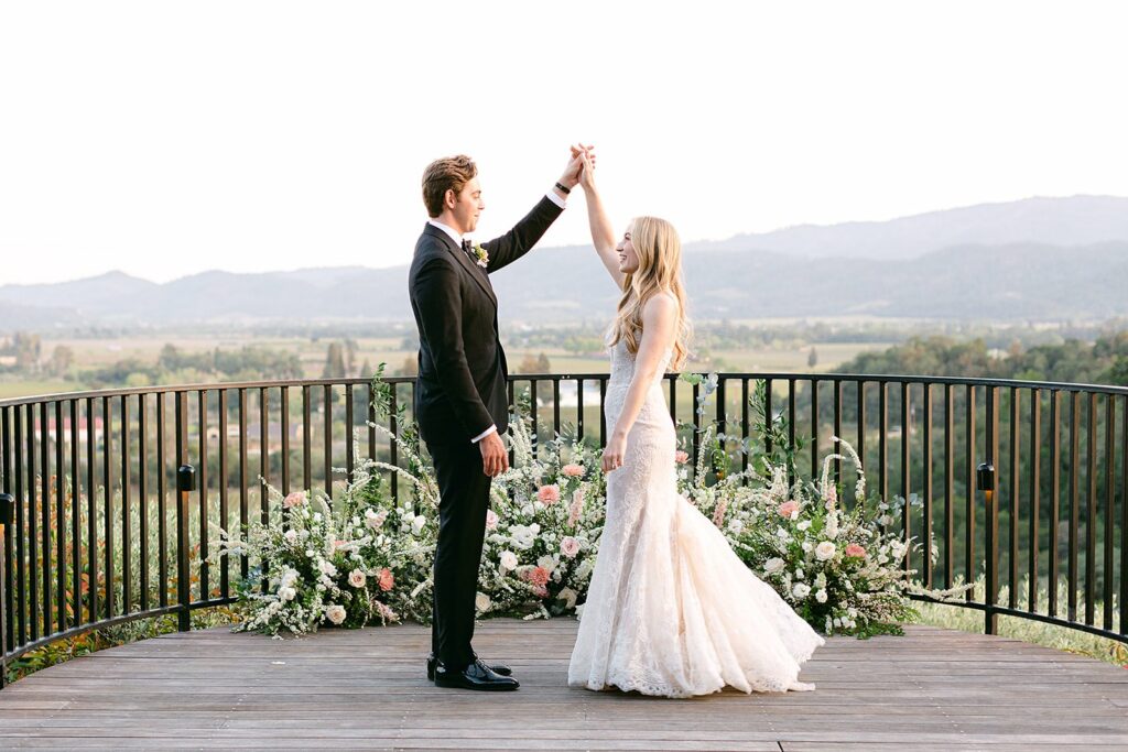 A bride and groom stand on a wooden deck surrounded by floral arrangements, with the groom lifting the bride's hand. A scenic landscape with mountains and valleys is visible in the background.