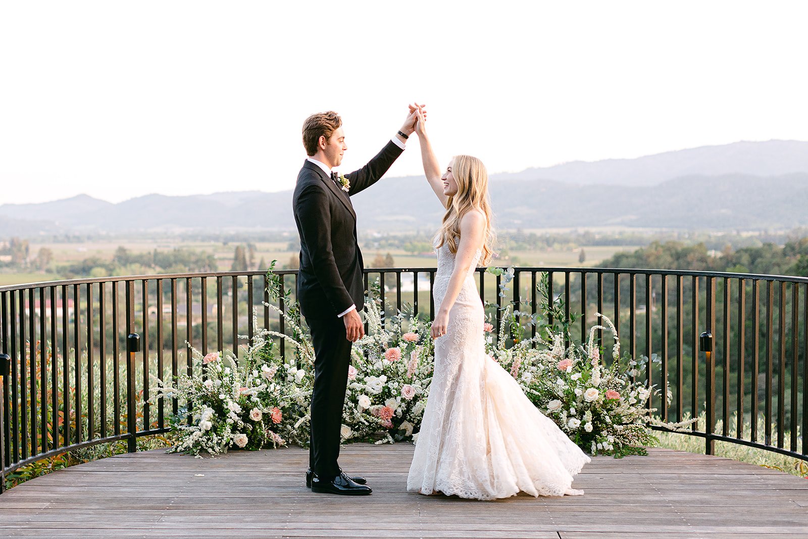 A bride and groom stand on a wooden deck surrounded by floral arrangements, with the groom lifting the bride's hand. A scenic landscape with mountains and valleys is visible in the background.